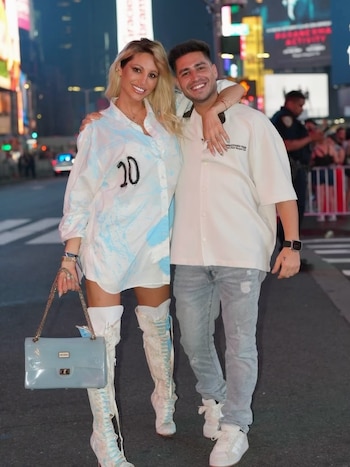 Una mujer y un hombre posan en una calle urbana iluminada. Ella viste camisa blanca con el número 20 y botas altas. Él lleva camisa clara, jeans y zapatillas