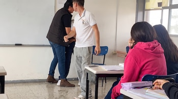 Dos hombres de pie frente a un pizarrón blanco en un aula, con tres estudiantes sentados en pupitres al frente y una ventana a la derecha.