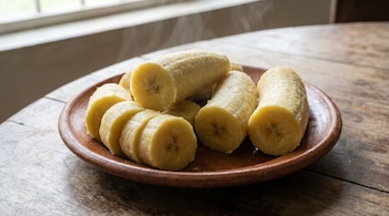 Plátano macho cocido, algunos enteros y otros en rodajas, humeando en un plato de barro sobre una mesa de madera. Una ventana está al fondo.