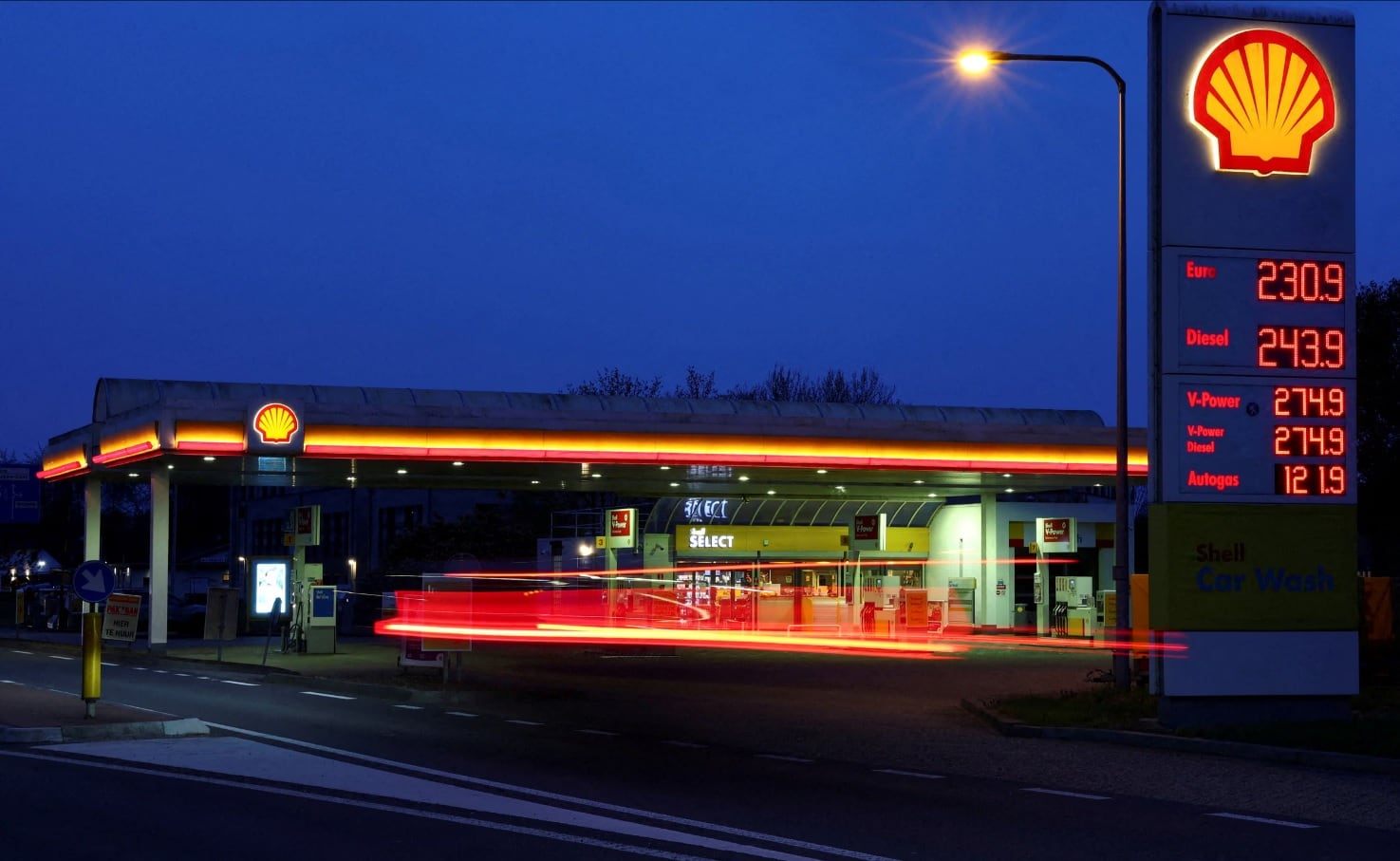 Una estación de servicio Shell se ilumina por la noche con un poste de precios digital mostrando el coste de varios tipos de combustible, mientras rastros de luz de vehículos crean dinamismo en la imagen.