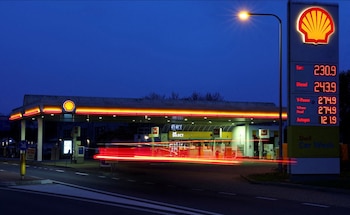 Vista nocturna de una gasolinera Shell con su logotipo brillante y un gran letrero digital de precios de combustible. Rastros de luz roja de vehículos se ven en primer plano