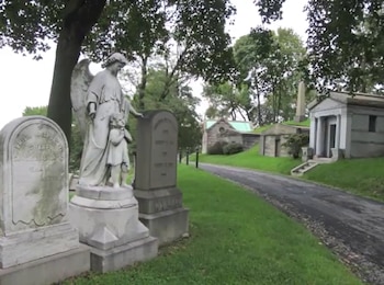 Una estatua de un ángel y un niño junto a lápidas en un cementerio. Un sendero serpentea a través del césped con mausoleos y árboles en el fondo