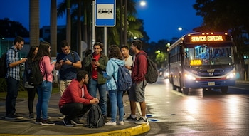 Ocho estudiantes con mochilas esperan en una parada de autobús iluminada por la noche, bajo un letrero de autobús. Un bus se acerca por la calle mojada.