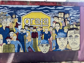 Un mural colorido en una pared exterior muestra una gran cantidad de personas con estilo de dibujos animados y el letrero amarillo 'grafa' en el centro
