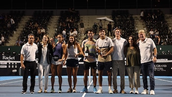 Nueve personas posan de pie junto a la red de una cancha de tenis nocturna mientras sostienen trofeos y botellas ante el público