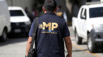 Vista trasera de un hombre con un chaleco azul oscuro del Ministerio Público, logo dorado "MP", y una mochila negra. Vehículos blancos borrosos al fondo.