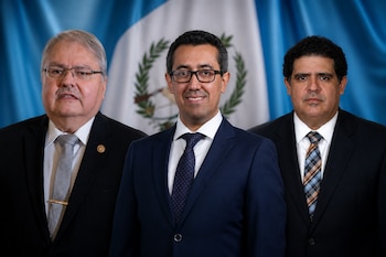 Tres hombres formales vistiendo traje y corbata posan frente a la bandera azul y blanca de Guatemala.