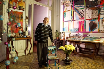 Un hombre en suéter a rayas, pantalón oscuro, sostiene una silla pintada. Hay globos decorativos, cuadros abstractos, mesa de madera, jarrón con flores amarillas