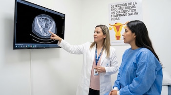 Doctora en bata blanca señala tomografía computarizada abdominal en monitor a paciente en bata azul. Póster sobre detección de endometriosis al fondo.