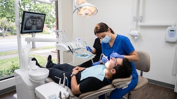 Hombre en silla dental, dentista y asistente, equipos odontológicos, monitor con radiografía dental, ventana exterior.
