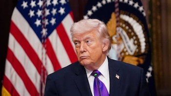 Primer plano del Presidente Donald Trump con traje oscuro y corbata morada, mirando a la izquierda, con la bandera de EE. UU. y el sello presidencial de fondo