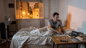 Hombre sudoroso sentado en cama desordenada, junto a mesa con reloj digital 03:45, teléfono, libros y vasos. Ventilador y ventana abierta.