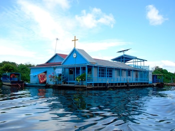 Estructura flotante de madera azul con una cruz amarilla en el techo, situada sobre el agua junto a botes y vegetación