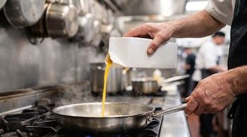 Manos vierten huevo líquido desde un cartón blanco hacia una cacerola de acero inoxidable en una estufa de cocina profesional, con vapor.
