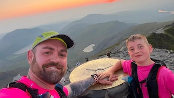 Un hombre con gorra y un niño con camiseta rosa apoyan sus manos sobre un disco dorado con relieve en la cima de una montaña