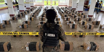 Vista trasera de un militar de Marina frente a numerosas pilas de paquetes envueltos en una sala con cinta de "Escena del Crimen" en el suelo