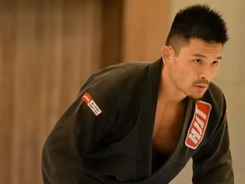 Hombre con uniforme negro de artes marciales y parches bordados, inclinado hacia adelante con mirada concentrada
