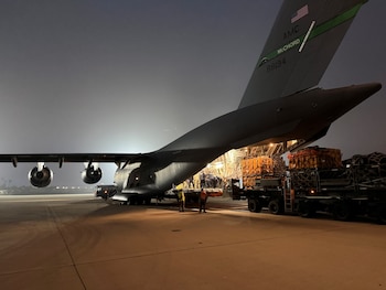 Avión de transporte militar gris en pista de aeropuerto por la noche, con rampa de carga trasera abierta. Varias personas y un camión cargan palés en la aeronave
