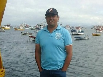 Hombre de mediana edad con gorra negra y camiseta polo azul, jeans, sonríe a la cámara, de pie frente a un puerto con numerosos barcos pesqueros