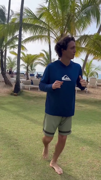 Un hombre descalzo camina sobre el césped cerca de palmeras y reposeras de playa