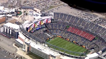 Vista aérea de un gran estadio de fútbol con gradas llenas y un campo verde. Hay edificios adyacentes y grandes estacionamientos con coches