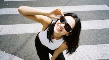 Vista aérea de una joven sonriente con cabello oscuro y gafas de sol, vistiendo una camiseta blanca y pantalones negros, posando sobre un paso de cebra.