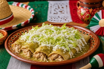 Enchiladas de pollo en salsa verde con lechuga y queso sobre un plato de barro, junto a un sombrero de paja y adornos con colores patrios.