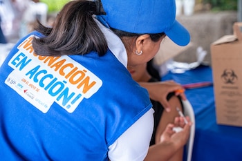 Trabajadora de la salud con chaleco azul y gorra administra una vacuna en el brazo de una persona. El chaleco dice "Vacunación en Acción". Al fondo, un recipiente de riesgo biológico