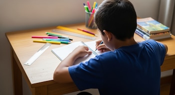Niño latino de espaldas escribiendo en un cuaderno con un lápiz. En el escritorio de madera hay útiles escolares, libros y luz natural ilumina la escena.