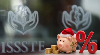Una alcancía con gorro navideño, monedas apiladas y un símbolo de porcentaje rojo ante un cristal con el logotipo grabado del ISSSTE.