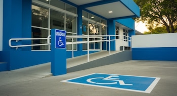 Fachada azul de un edificio con puertas automáticas de vidrio, rampa de acceso, barandales blancos, un cartel y un símbolo pintado de acceso universal.