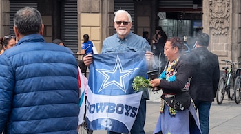 Un hombre de cabello blanco sostiene una bandera azul de Dallas Cowboys junto a una mujer que lleva plantas y un recipiente de humo.