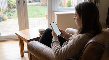 Una mujer sentada en un sillón de cuero mira un teléfono móvil con texto en pantalla, junto a una mesa de madera y una ventana con jardín.