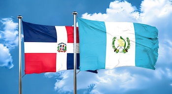 Dos banderas ondean en mástiles plateados contra un cielo azul claro con nubes. A la izquierda, la bandera dominicana; a la derecha, la bandera guatemalteca.