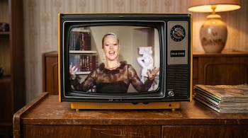Un televisor retro amarillo y negro con la imagen de Pinky en pantalla, sonriente y con los brazos abiertos, posado sobre un mueble de madera con revistas a un lado.