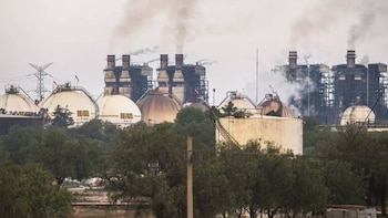 Tanques esféricos de almacenamiento blanco y torres industriales con columnas de humo detrás de varios árboles