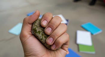 Primer plano de una mano adolescente de piel morena clara, tensa y con suciedad, apretando una piedra grande y cubierta de musgo, con cuadernos desenfocados en el suelo.