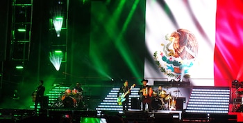 Músicos tocan sus instrumentos en un escenario iluminado frente a una pantalla con la bandera de México
