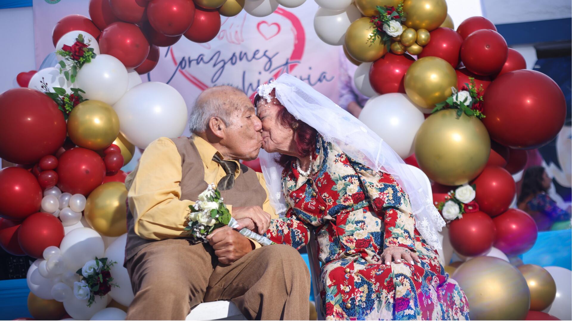 Una pareja de ancianos comparte un tierno beso, celebrando su amor eterno bajo un arco festivo de globos rojos, blancos y dorados.