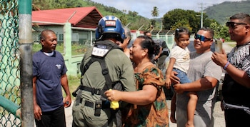 Un miembro de la Guardia Costera de EE. UU. con casco azul interactúa con una mujer. Hombres y una niña observan. Detrás hay una valla, casas y vegetación