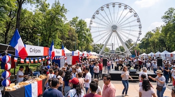 Personas recorren puestos decorados con banderas francesas frente a músicos y una gran rueda de la fortuna al fondo en un parque al aire libre.