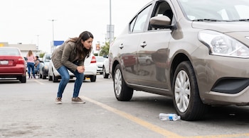 Una mujer inclinada observa la parte baja de un automóvil junto a una botella de plástico tirada cerca de una llanta en un estacionamiento.