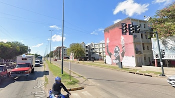 Vista callejera con un hombre en moto en primer plano, vehículos en una avenida, y edificios con un gran mural de Lionel Messi bajo un cielo azul con nubes