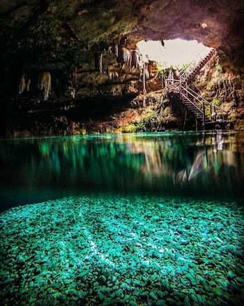Cueva con estalactitas, una abertura de luz y escaleras de madera. Agua turquesa con reflejos en la superficie. Fondo subacuático cubierto de piedras claras