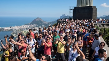 Multitud de turistas en la plataforma del Cristo Redentor, muchos tomando fotos. Al fondo, la ciudad de Río de Janeiro, el océano y el Pan de Azúcar.