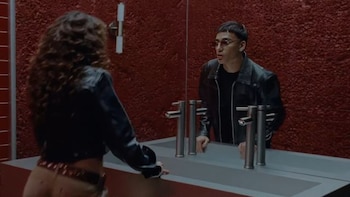 Reflejo de un hombre con gafas de sol, chaqueta oscura y camiseta negra; persona de espaldas con cabello rizado; lavabo blanco con dos grifos; pared roja
