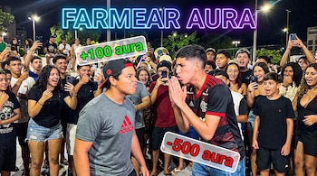 Dos jóvenes en primer plano se enfrentan rodeados por una multitud, algunos con teléfonos móviles. La imagen tiene texto neón "FARMEAR AURA" y stickers "+100 aura" y "-500 aura".
