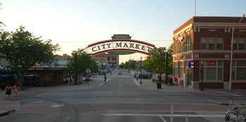 Un letrero arqueado de 'Historic City Market Kansas City' sobre una calle con edificios de ladrillo, autos estacionados, árboles, peatones y postes de luz