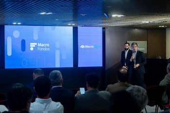 Dos hombres en traje se encuentran junto a una pantalla azul con las inscripciones Macro Fondos y Macro Agro frente a un público de espalda