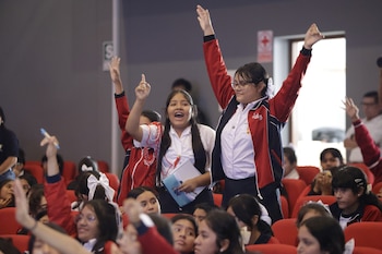 Grupo de jóvenes estudiantes con uniformes de colegio, sentadas en butacas rojas en un auditorio, levantan las manos con entusiasmo